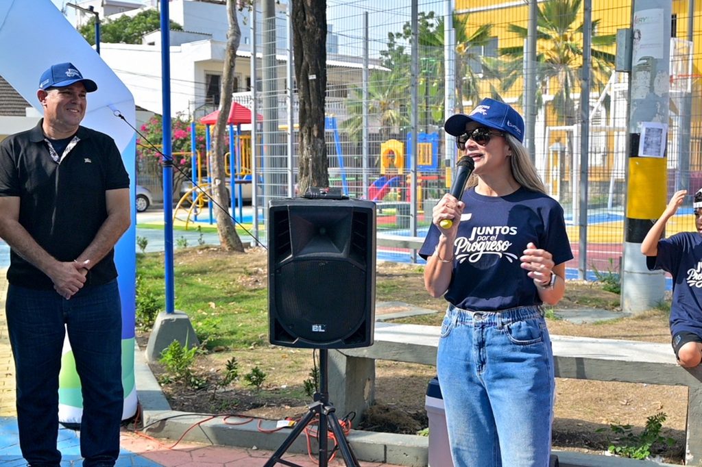 Mujer con gorra azul y camiseta "Juntos por el Progreso" habla por micrófono junto a un hombre, en el parque del barrio Torices, Cartagena.